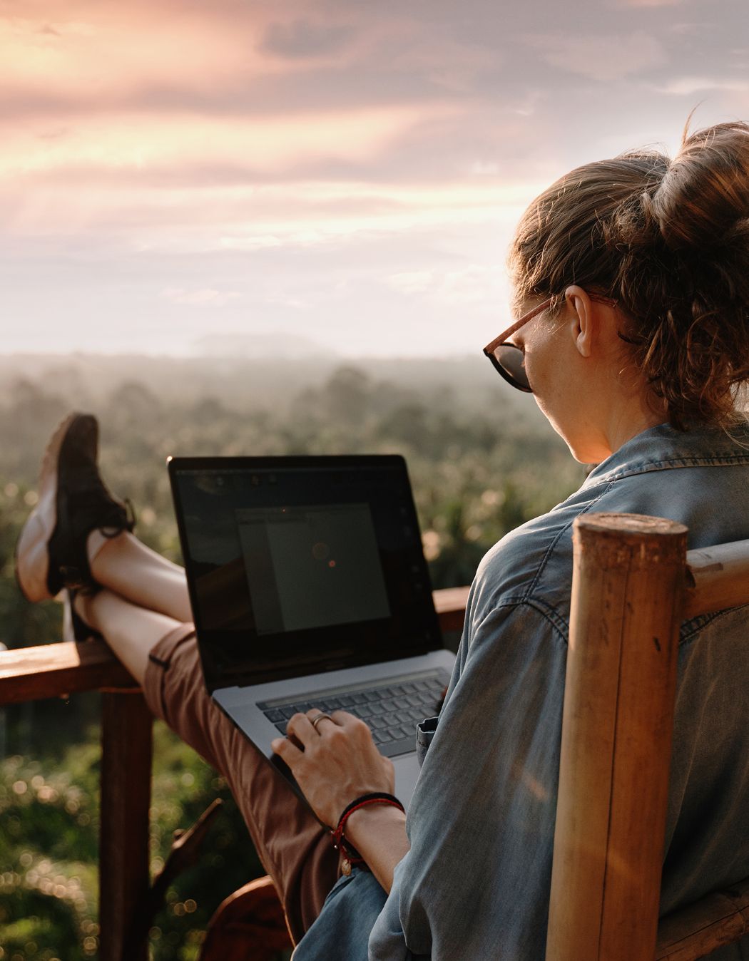A woman works on a laptop, there is a forest and sunset in the distance. For Blog: Certified Financial Planner Designation